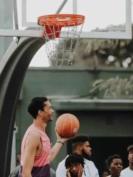 Tall basketball player holding the ball underneathe the basket while other players watch him